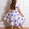 Une jeune fille vue de dos, portant une robe blanche à imprimés de tulipes mauves, avec une fleur assortie dans ses cheveux ondulés.