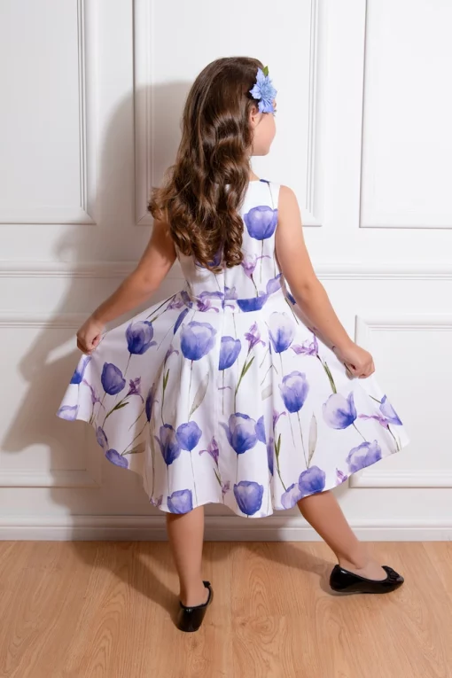Une jeune fille vue de dos, portant une robe blanche à imprimés de tulipes mauves, avec une fleur assortie dans ses cheveux ondulés.