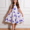 Une jeune fille souriante faisant face à la caméra, vêtue d'une robe blanche à imprimés de tulipes mauves, avec une fleur dans ses cheveux.