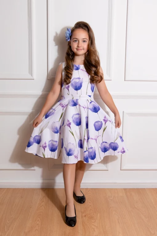 Une jeune fille souriante faisant face à la caméra, vêtue d'une robe blanche à imprimés de tulipes mauves, avec une fleur dans ses cheveux.