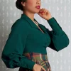 Femme en blouse verte et jupe tartan, regard contemplatif vers le côté.