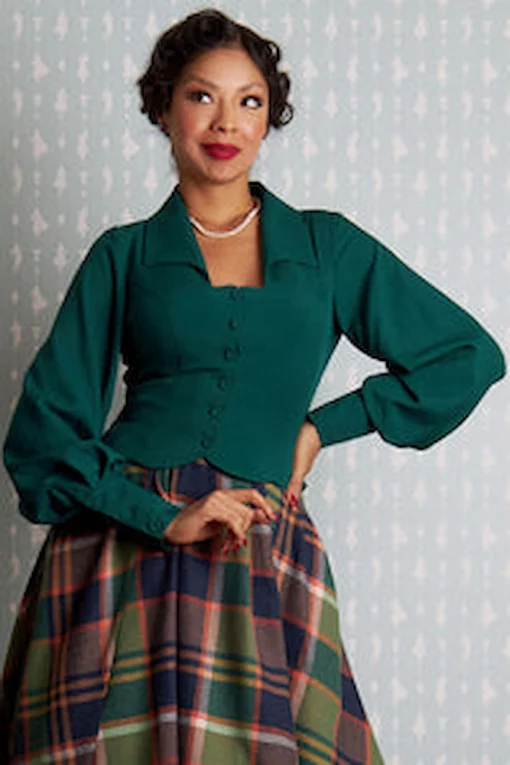 Femme avec une blouse verte à col et manches longues et une jupe à carreaux plissée, posant avec assurance.